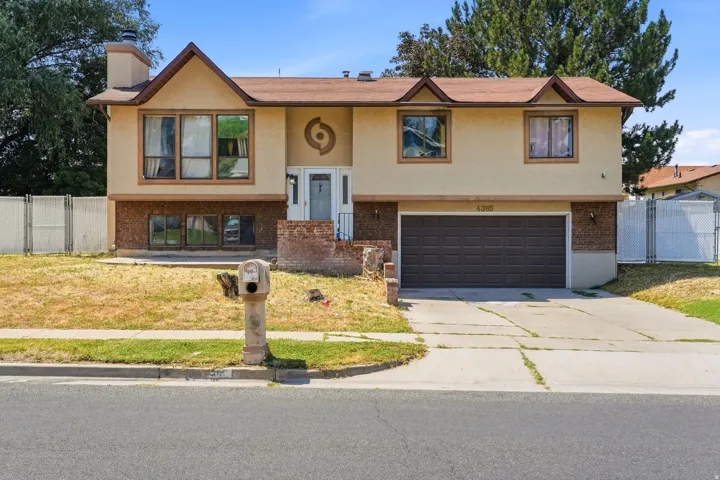 Split-level home featuring a tan and brick exterior with multiple gabled rooflines, a two-car garage with dark paneled doors, and a manicured front lawn bordered by mature landscaping and white vinyl fencing