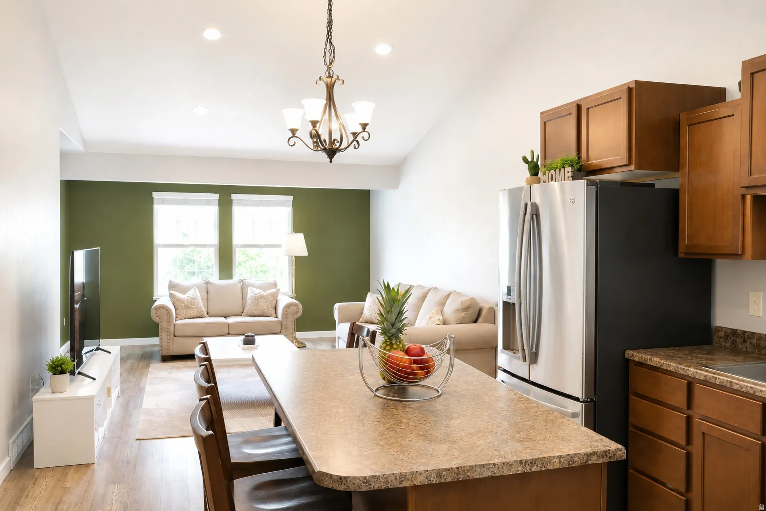 Open-concept living area featuring a vaulted ceiling with recessed lighting and a chandelier, wood-finish flooring, a kitchen island with seating, stainless steel refrigerator, and brown cabinetry with a speckled countertop