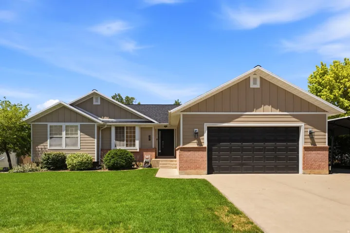 Front exterior with tan siding and brick accents, featuring a two-car garage, a well-maintained lawn, and established landscaping