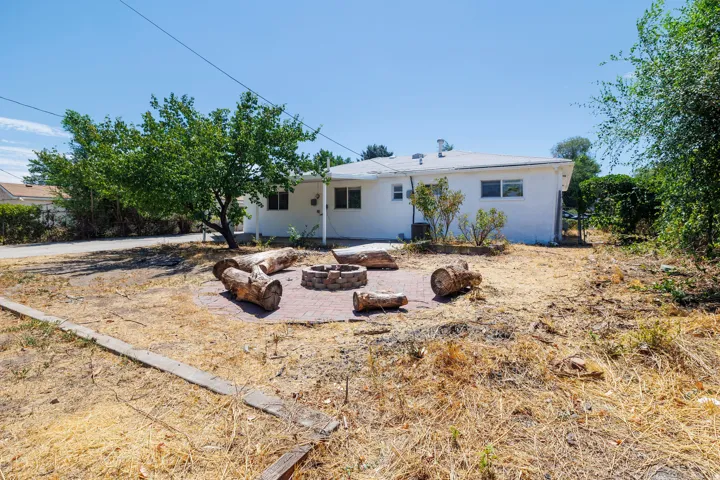 Expansive backyard featuring a brick paver patio with a built-in fire pit, mature tree providing shade, and a white exterior residence with multiple windows