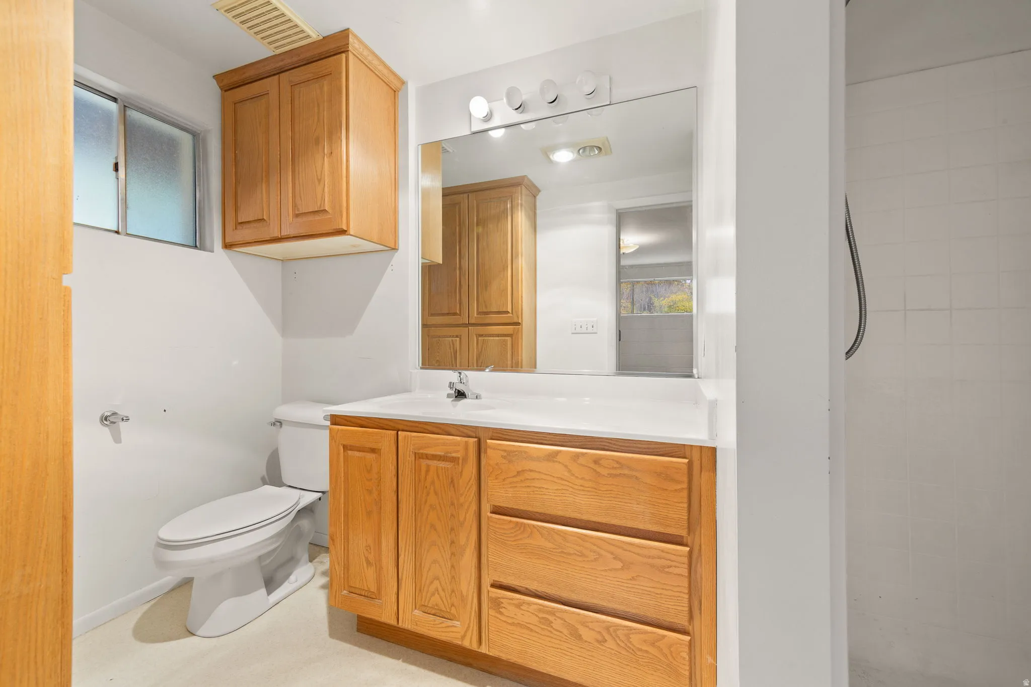 Bathroom featuring a light wood-finish vanity with an integrated sink and mirror, a white toilet, an overhead wood-finish cabinet, and a tiled shower stall