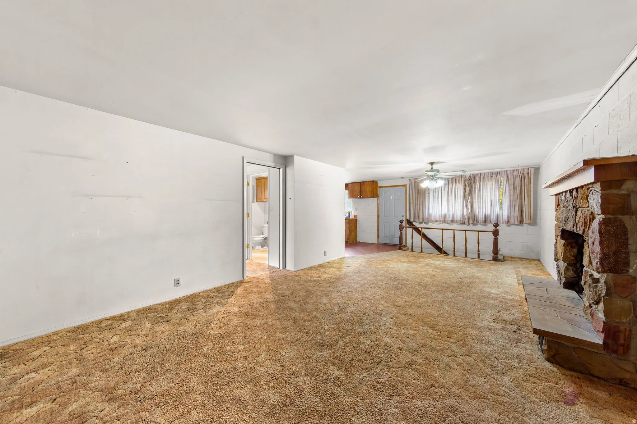Spacious interior featuring a stone fireplace with a wood mantle, textured carpeting, a wood-finish handrail, and an overhead ceiling fan