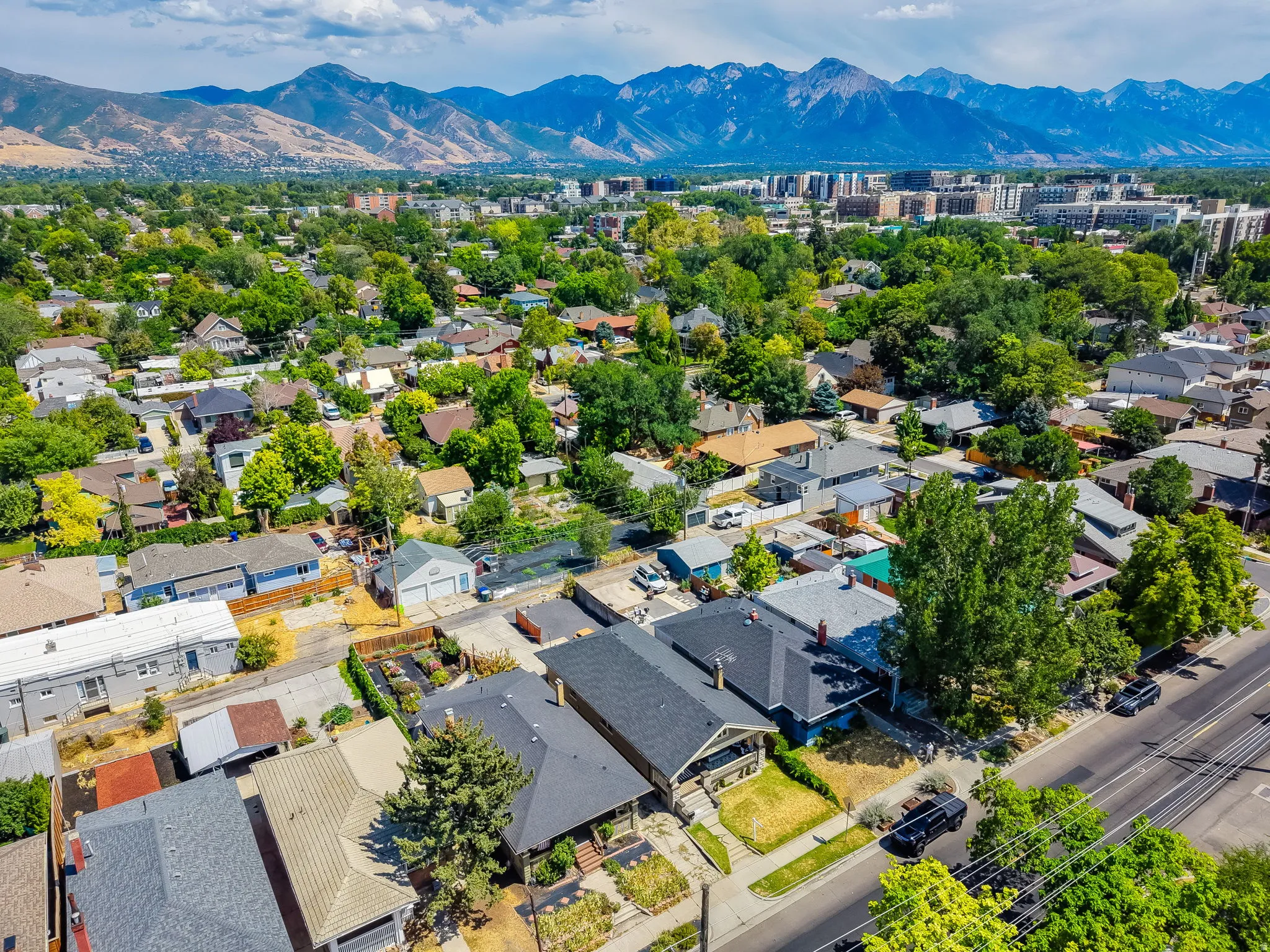 Aerial view showcasing this residential property nestled within a tree-lined neighborhood, featuring a dark-roofed home with defined architectural lines, mature landscaping throughout the lot, and proximity to the Wasatch mountain range