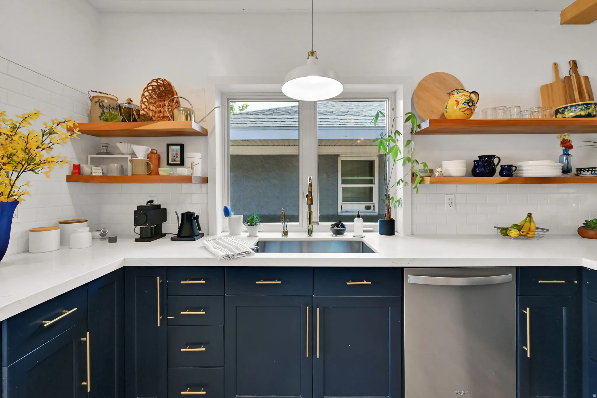 Kitchen featuring white subway tile backsplash, white countertops, deep blue lower cabinetry with brass-finish hardware, open wood-finish shelving, and a stainless steel dishwasher