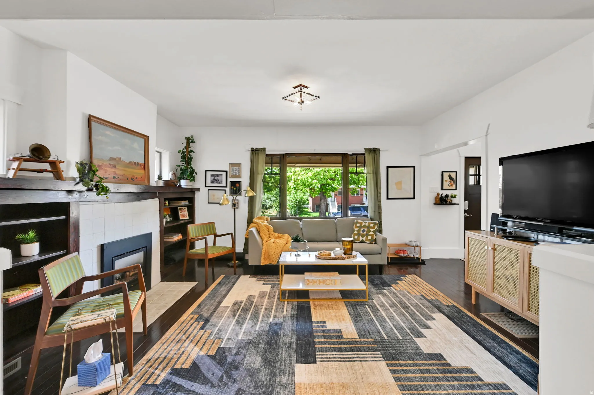 Spacious living area featuring dark wood-finish flooring, a central fireplace with built-in shelving, white tiled surround, and large front-facing windows