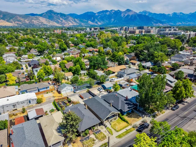 Aerial view showcasing this residential property nestled within a tree-lined neighborhood, featuring a dark-roofed home with defined architectural lines, mature landscaping throughout the lot, and proximity to the Wasatch mountain range