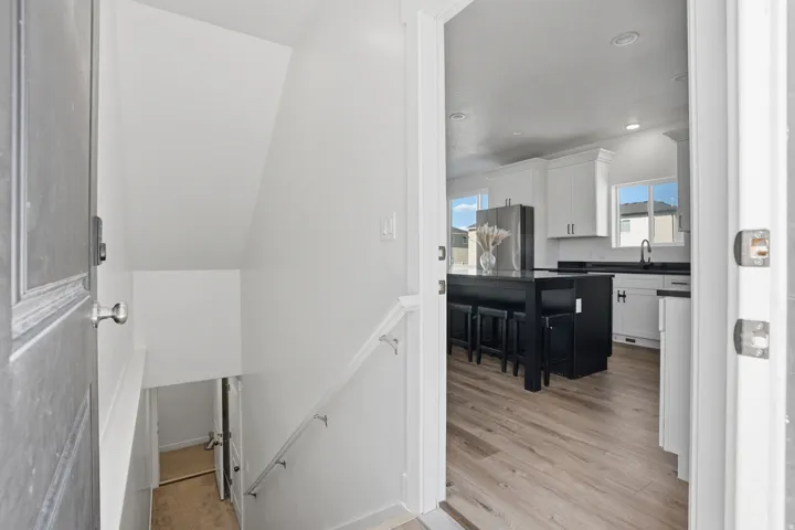 Inviting entryway featuring a dark-paneled door, light-toned wood-finish flooring, a white staircase with a metal handrail, and a bright kitchen with white cabinetry, dark countertops, and a contrasting dark island