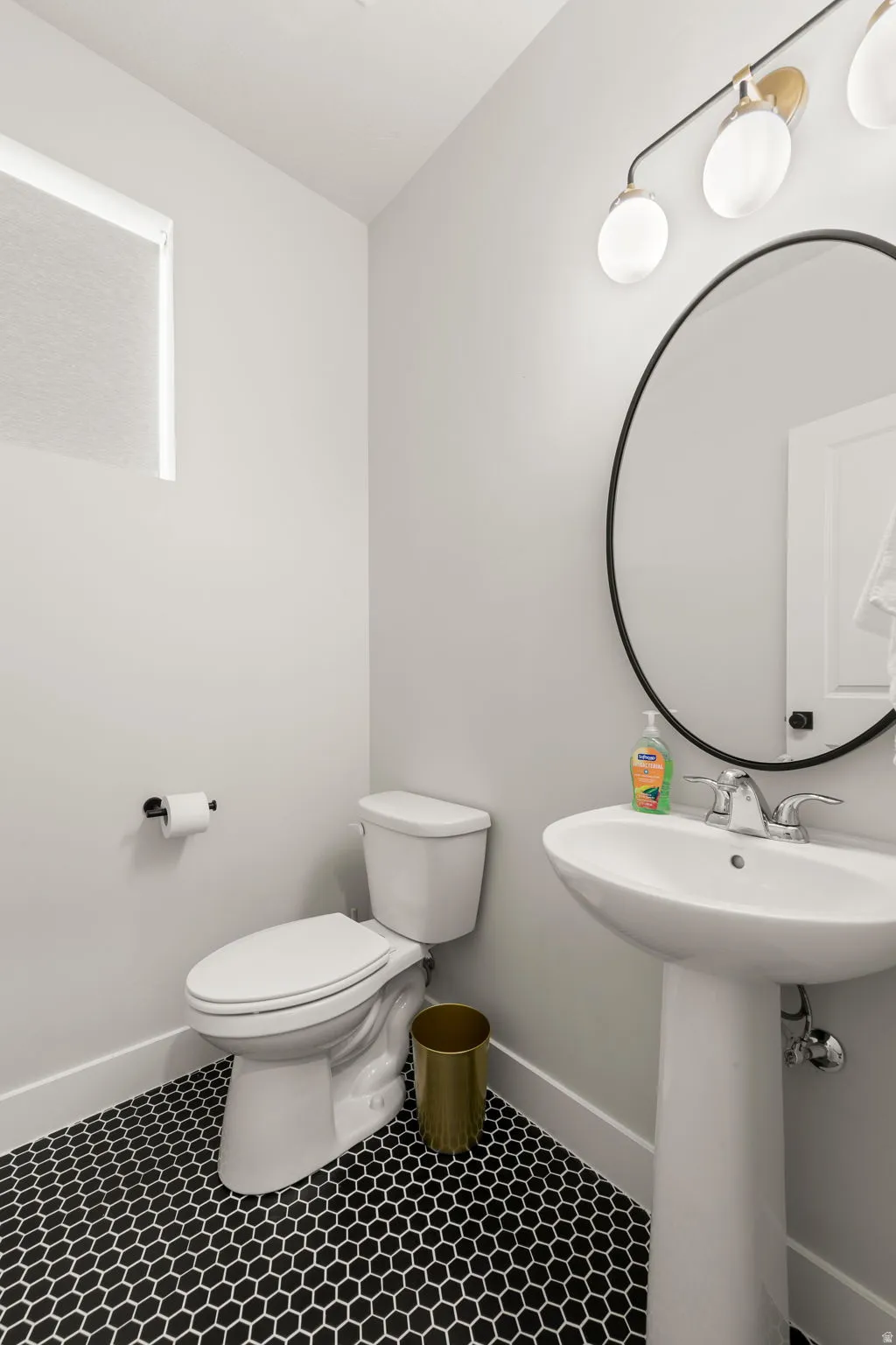 Pedestal sink with chrome faucet, complemented by a large circular mirror and a three-light vanity fixture