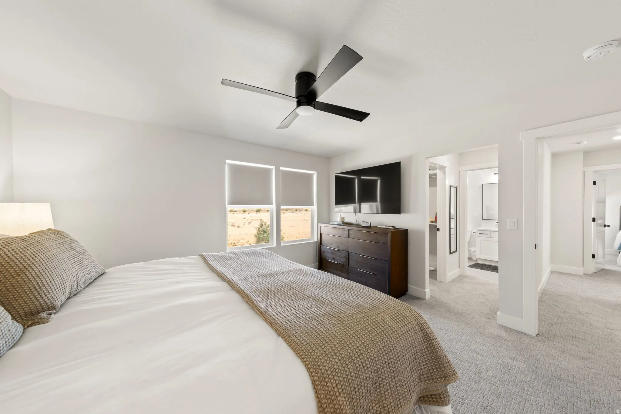 Spacious bedroom featuring light gray carpeting, a contemporary black ceiling fan, and two windows with roller shades