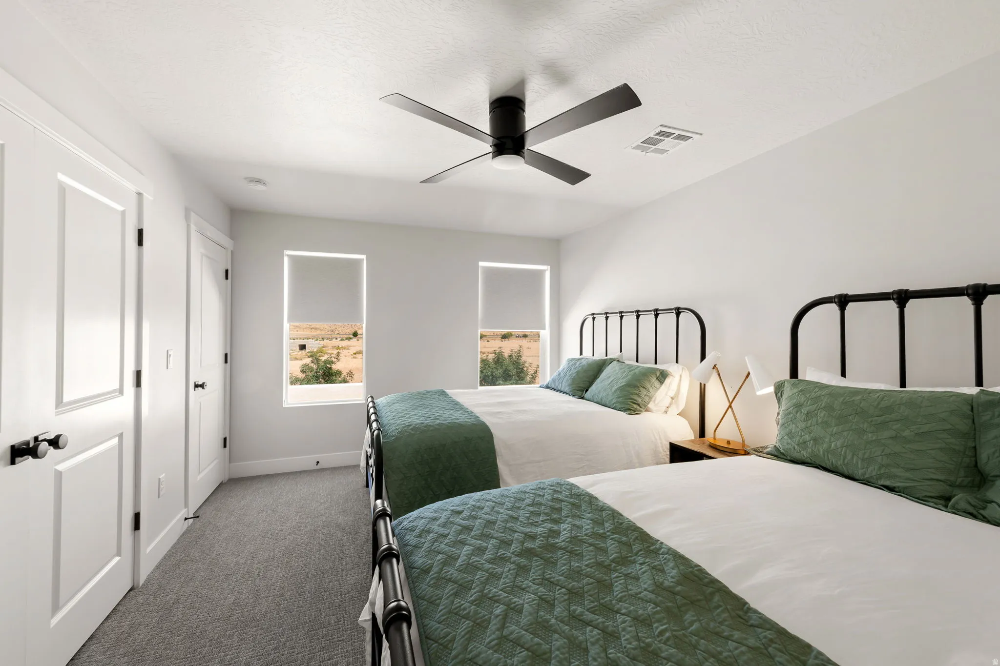 Bright bedroom featuring soft gray carpeting, white walls, two windows with roller shades, a black ceiling fan, and white panel doors with black hardware