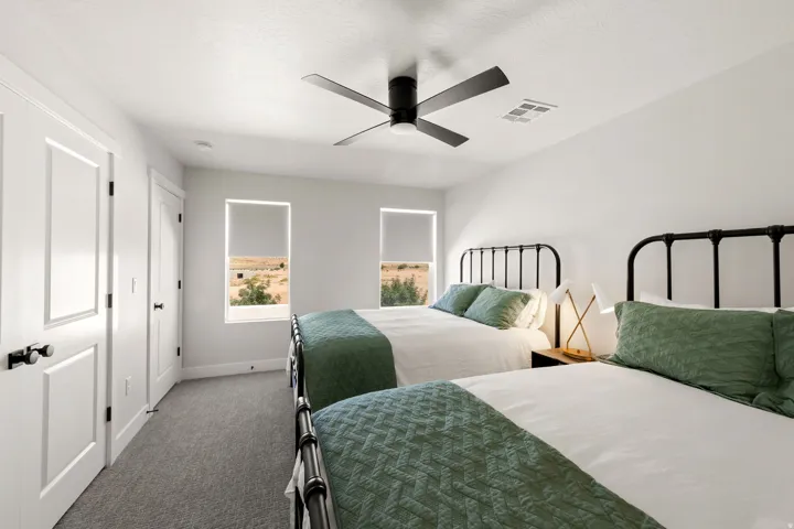 Bright bedroom featuring soft gray carpeting, white walls, two windows with roller shades, a black ceiling fan, and white panel doors with black hardware