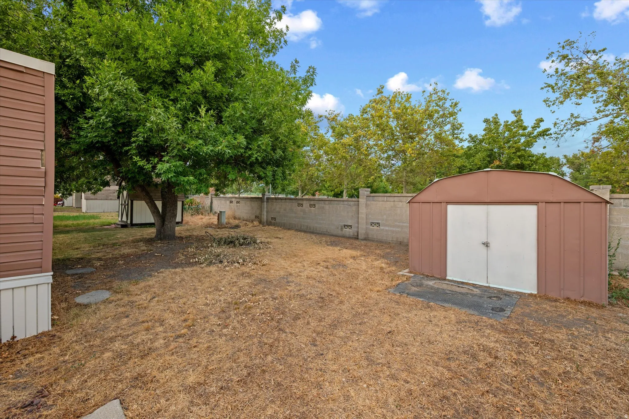 Rear yard featuring a brown metal storage shed with white double doors, a concrete block perimeter wall, and mature trees
