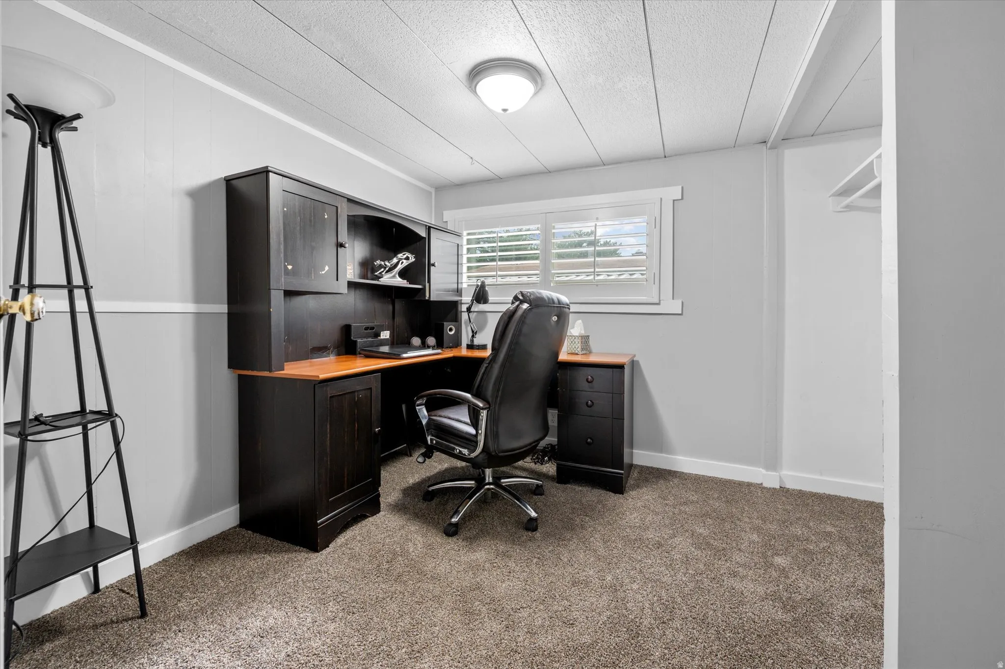 Carpeted room featuring a built-in dark wood desk with overhead cabinetry, a window with white louvered shutters, light gray wall paneling, and a white ceiling with recessed lighting