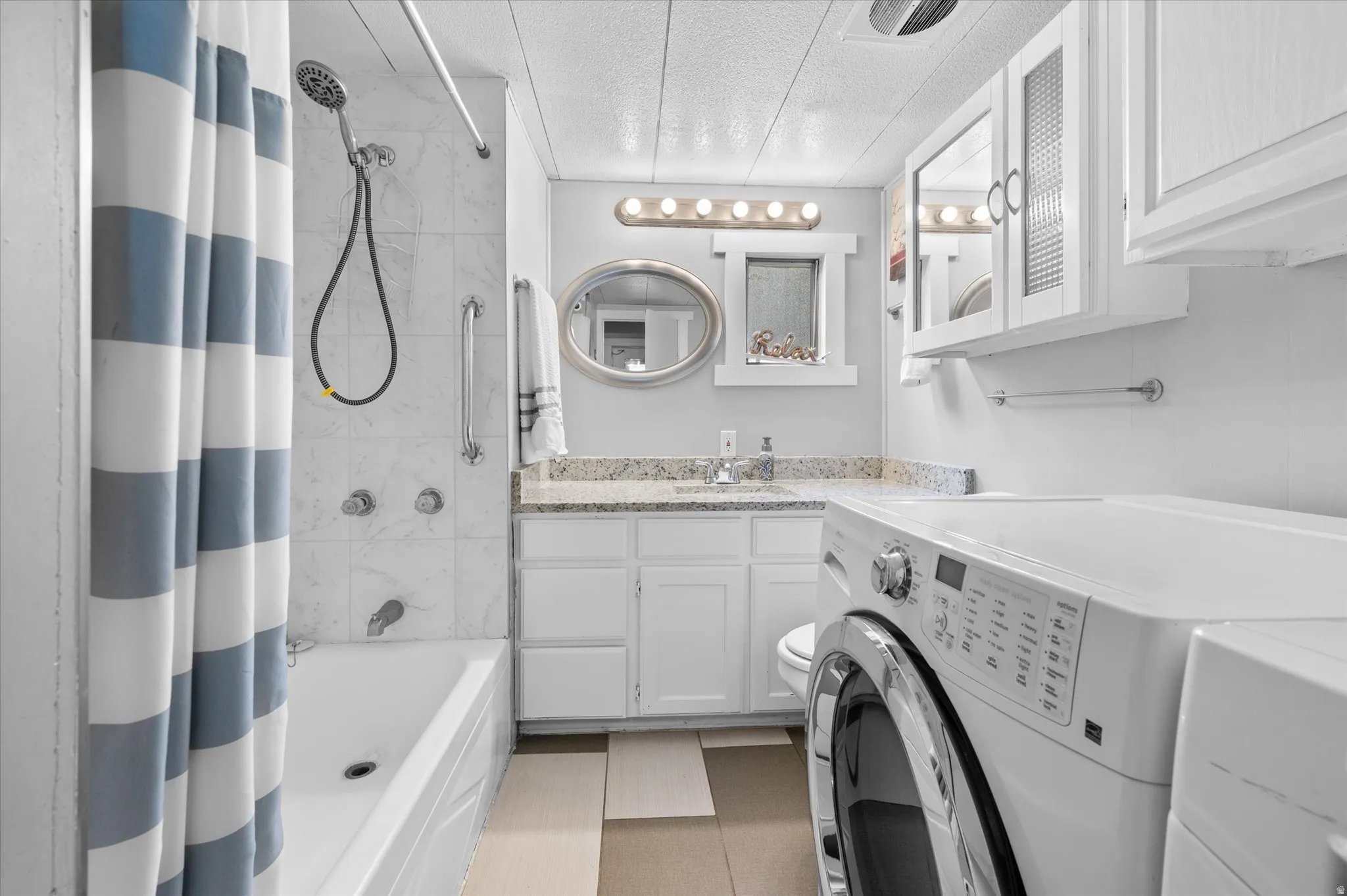 Bathroom featuring a bathtub with marble-look tile surround, a white vanity with a granite-look countertop, integrated sink, and upper cabinetry, and dedicated laundry appliance space