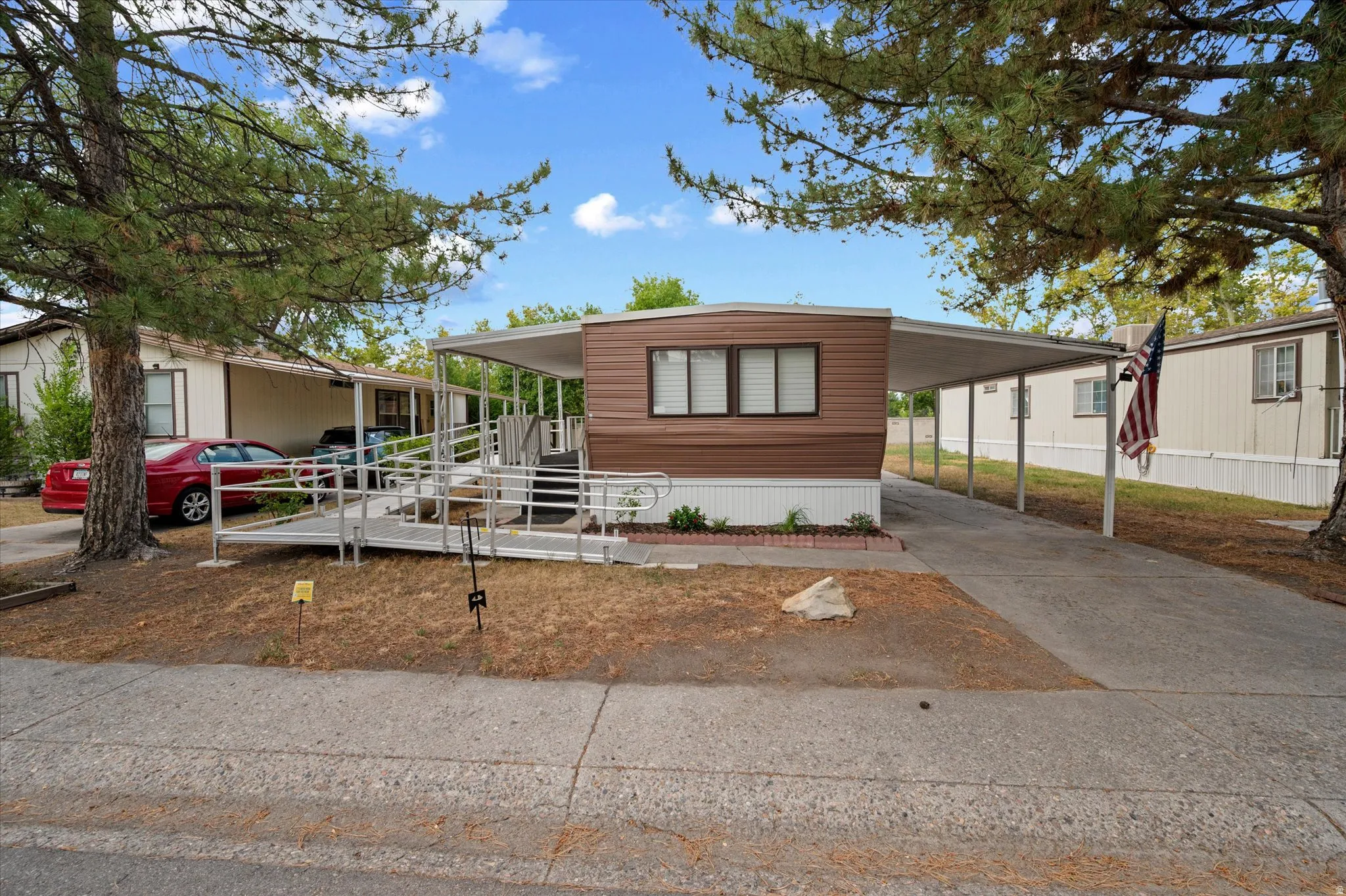 Brown-sided manufactured home featuring a covered carport, an accessible ramp, and mature trees providing shade