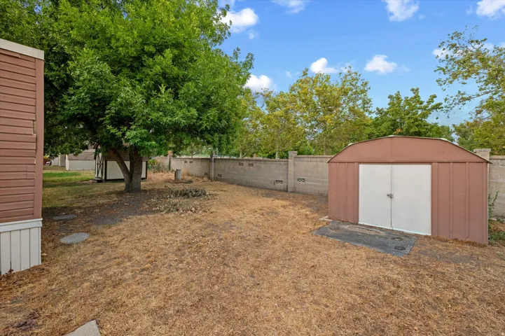 Rear yard featuring a brown metal storage shed with white double doors, a concrete block perimeter wall, and mature trees