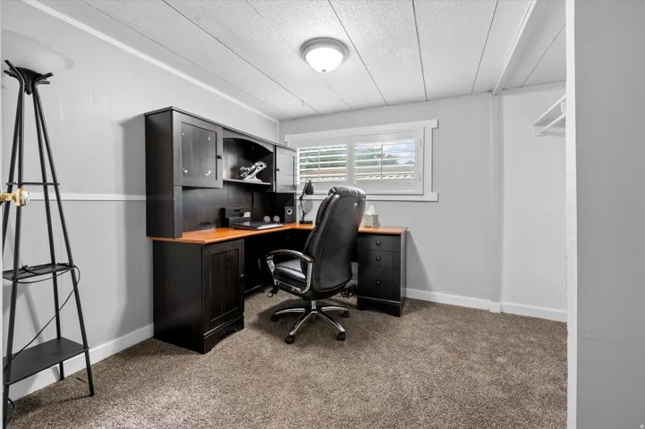 Carpeted room featuring a built-in dark wood desk with overhead cabinetry, a window with white louvered shutters, light gray wall paneling, and a white ceiling with recessed lighting