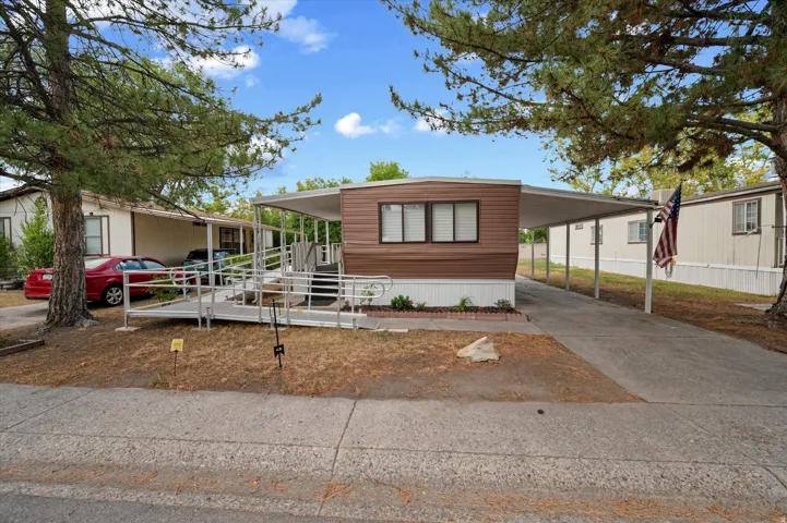 Brown-sided manufactured home featuring a covered carport, an accessible ramp, and mature trees providing shade