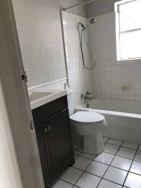 Bathroom featuring a white subway tile tub surround, integrated white bathtub, white floor tiles, a white toilet, and a dark wood-finish vanity with a white countertop