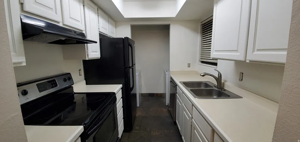 Galley kitchen featuring white cabinetry, light-toned countertops, a stainless steel double basin sink, and black appliances including a range and refrigerator