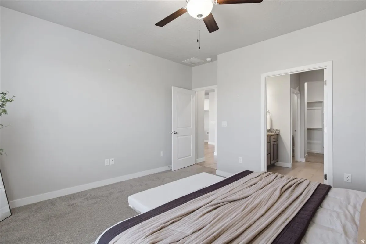 Carpeted bedroom featuring a ceiling fan with light fixture, light gray walls, white baseboards, and a doorway to a bathroom with a vanity and a separate closet with built-in shelving