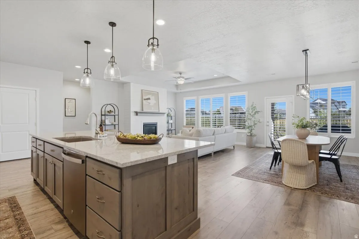 Open-concept living space featuring a spacious kitchen island with a built-in sink and pendant lighting, wood-finish flooring, a fireplace with a mantel, white window shutters, and a dining area with direct exterior access