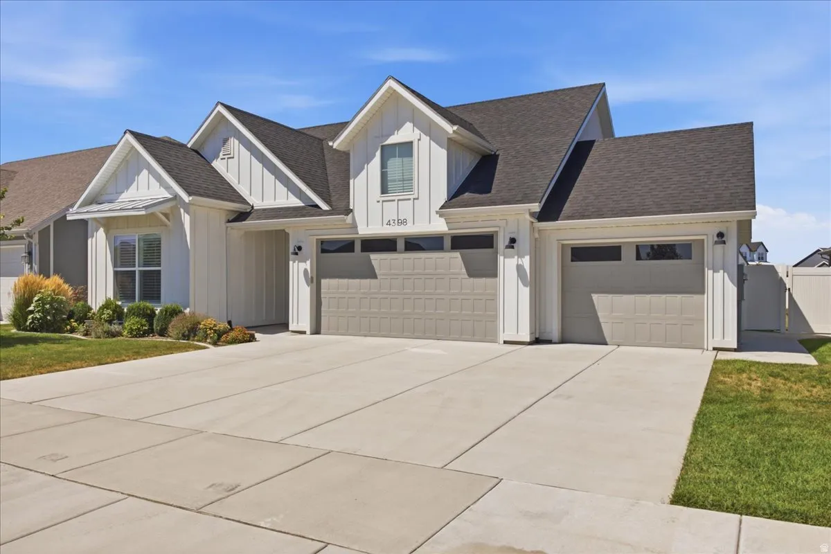 Modern farmhouse-style exterior featuring vertical white siding, dark shingle roof, three gray garage doors with window accents, a concrete driveway, and landscaped front yard