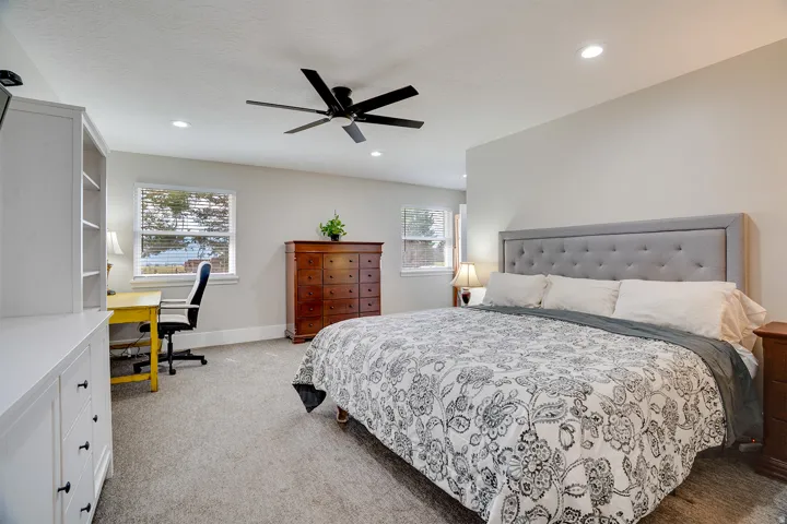 Spacious bedroom featuring light neutral wall tones, carpeted flooring, a ceiling fan, recessed lighting, and multiple windows with blinds