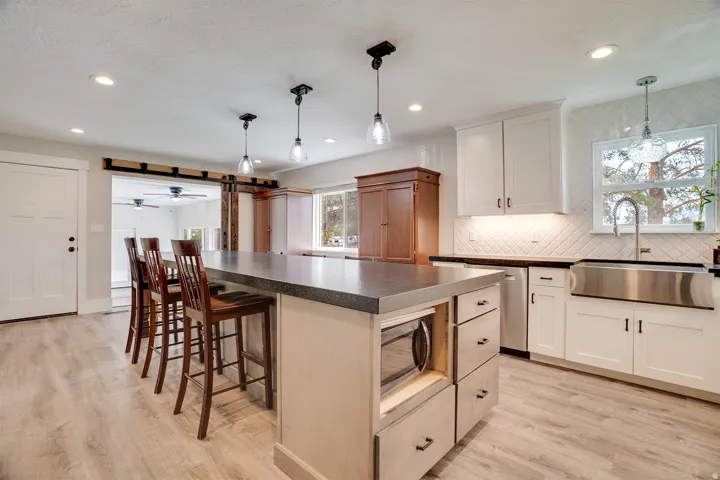 Spacious kitchen featuring a large island with built-in microwave, light wood-finish flooring, white shaker cabinetry with dark hardware, a stainless steel apron sink, and a white diagonal tile backsplash