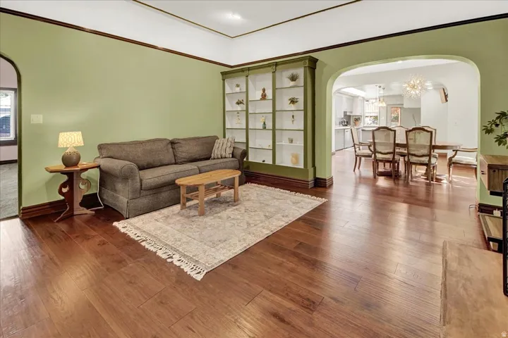 Living area featuring wood-finish flooring, built-in shelving, crown molding, and an arched doorway leading to a dining area