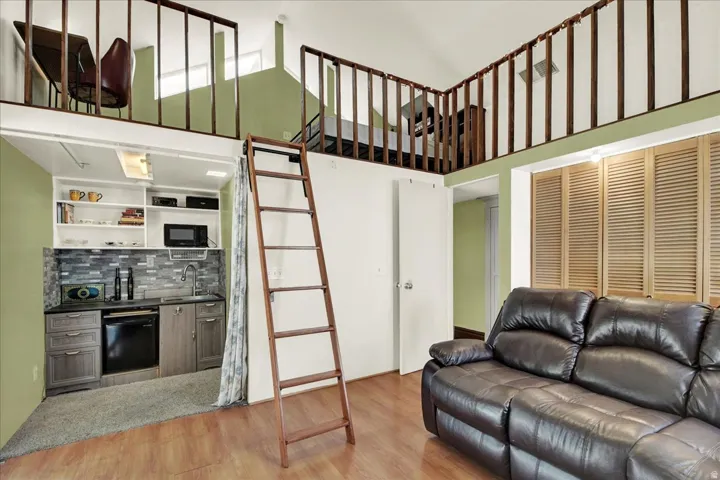 Bedroom 3 open-concept living space featuring wood-finish flooring, a built-in kitchenette with a tiled backsplash, and a lofted area with a wooden railing