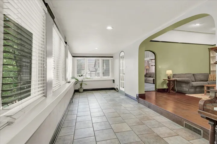 Sunlit room featuring multiple windows with blinds, tile flooring, and a recessed archway opening to an adjacent space with wood-finish flooring and sage green walls