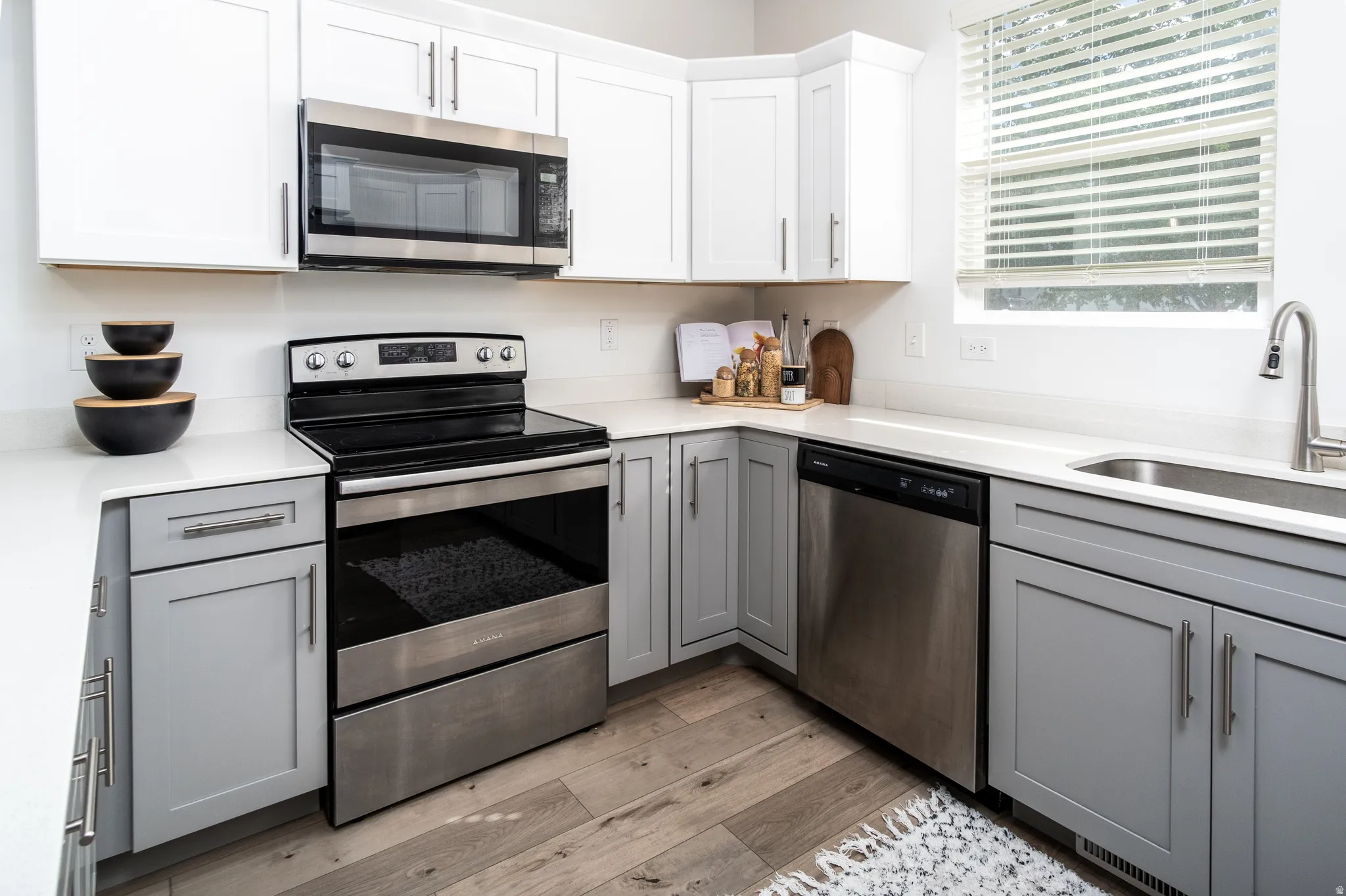 Kitchen featuring light wood-finish flooring, two-tone cabinetry with upper white shaker-style doors and lower gray shaker-style doors, light-toned countertops, and stainless steel appliances including a range, microwave, and dishwasher