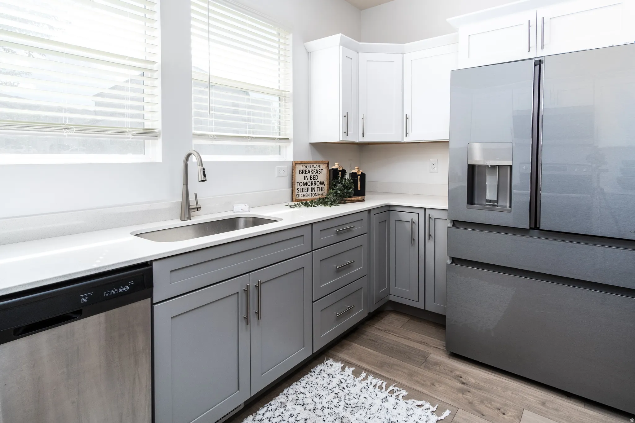 Contemporary kitchen featuring light gray base cabinetry, white upper cabinetry, light-toned countertops, a stainless steel sink with a gooseneck faucet, and integrated stainless steel appliances