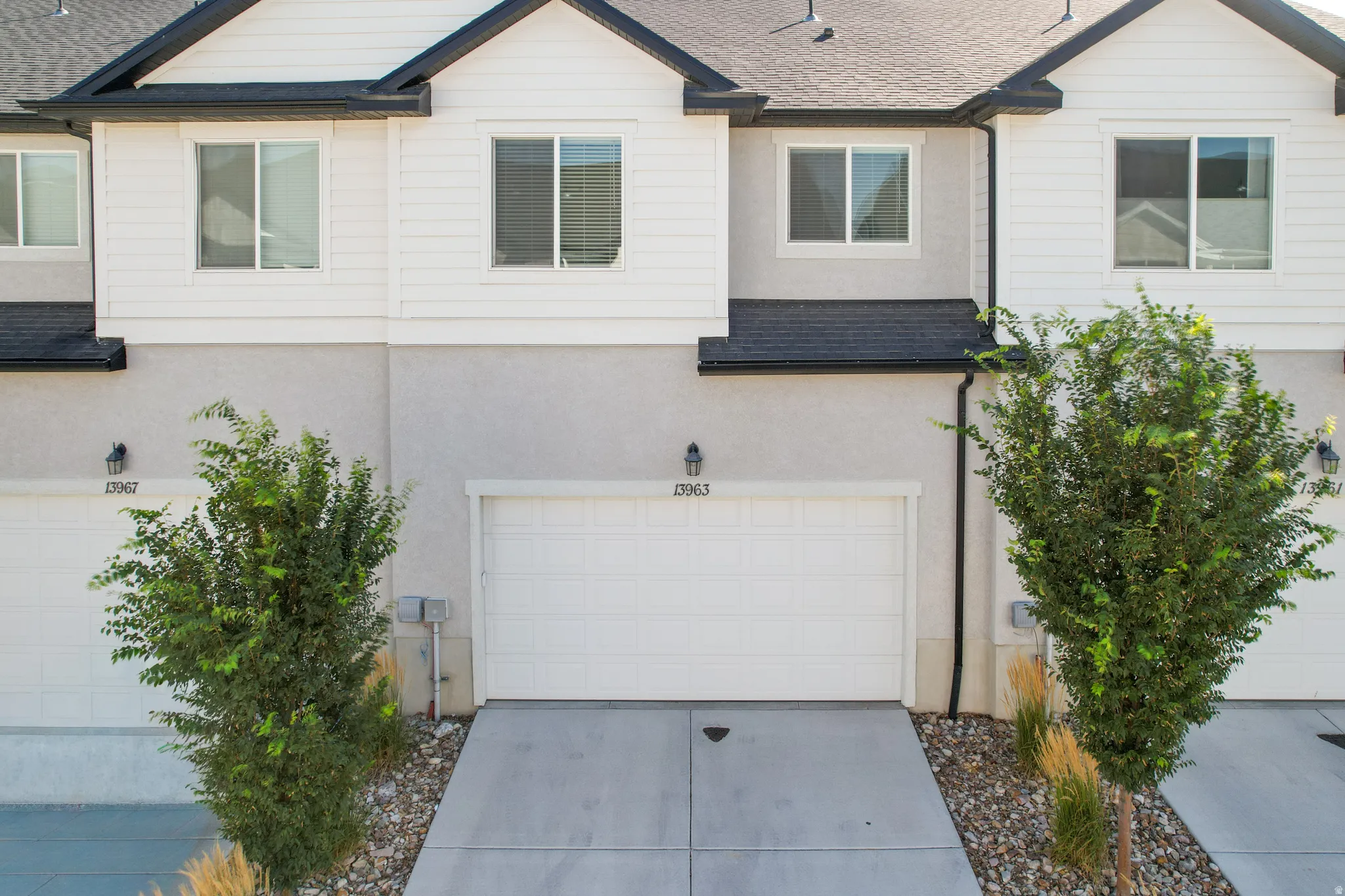 Contemporary townhome exterior featuring a white garage door, concrete driveway, decorative landscaping, and a two-tone facade with white siding and gray stucco