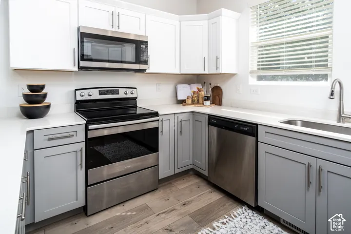 Kitchen featuring light wood-finish flooring, two-tone cabinetry with upper white shaker-style doors and lower gray shaker-style doors, light-toned countertops, and stainless steel appliances including a range, microwave, and dishwasher