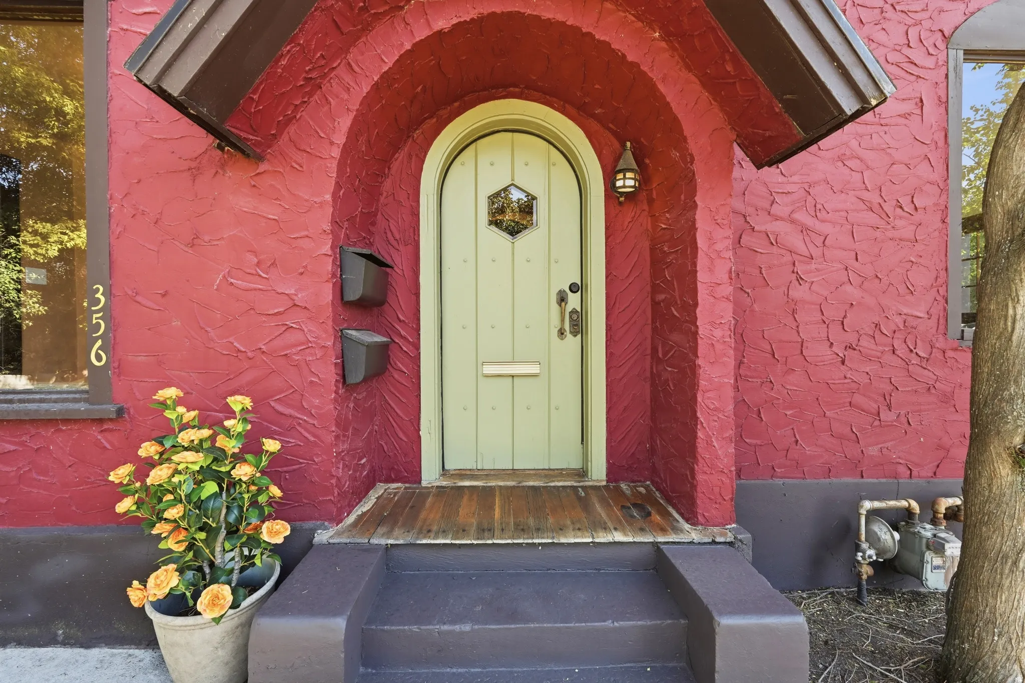 Arched red stucco entryway featuring a light green paneled door with decorative glass, a wall-mounted exterior light fixture, and a wood-finish porch landing