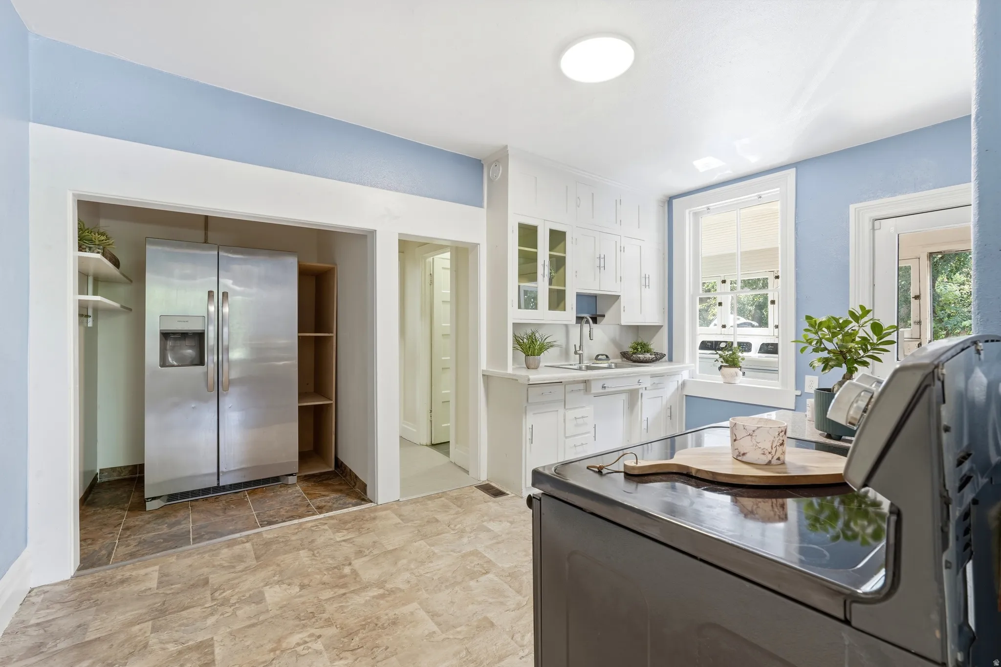 Kitchen featuring white cabinetry, a stainless steel side-by-side refrigerator, a white sink, light-toned flooring, and a large window with white trim