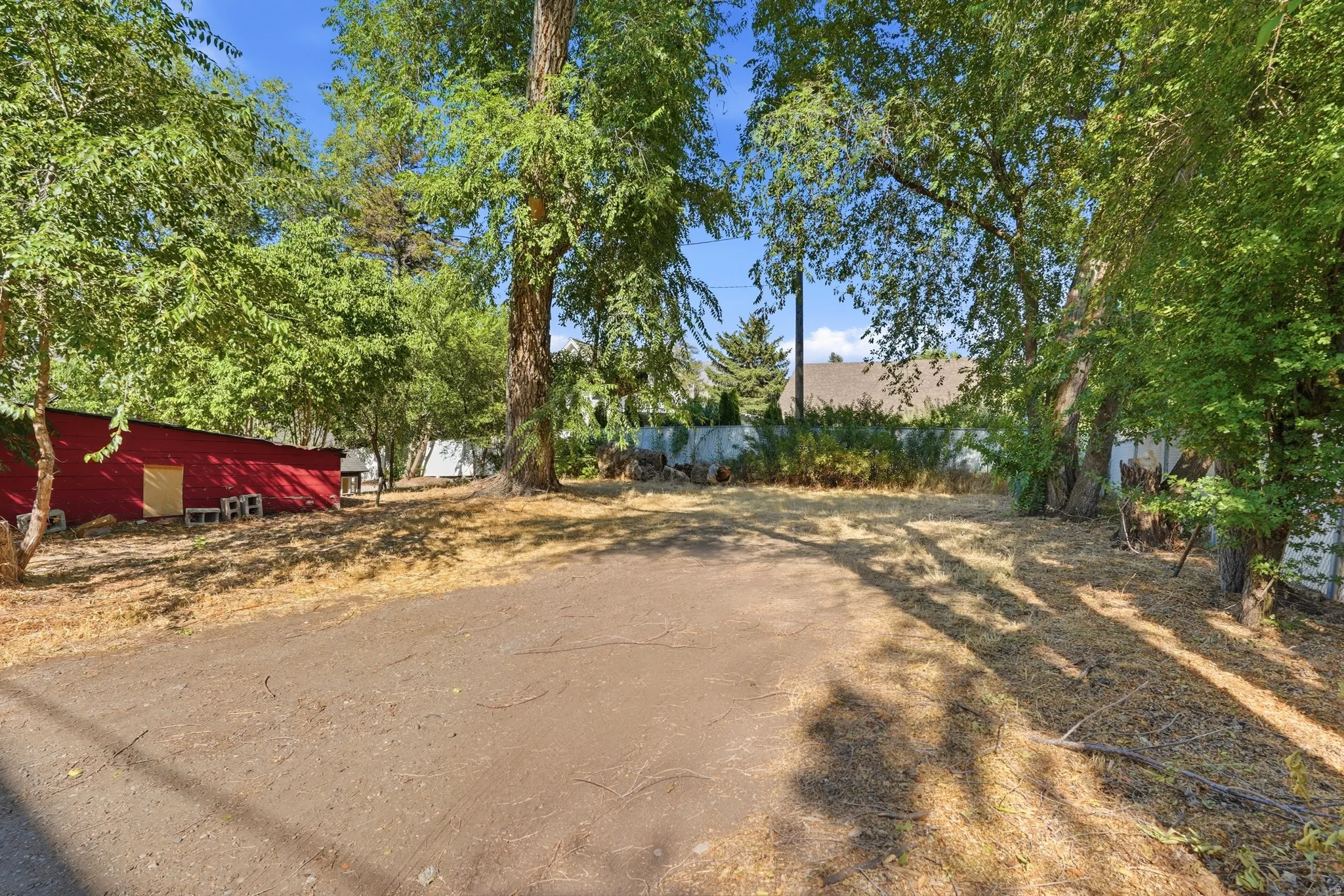 Spacious lot featuring mature trees providing ample shade, a red storage building, and a partially graveled area