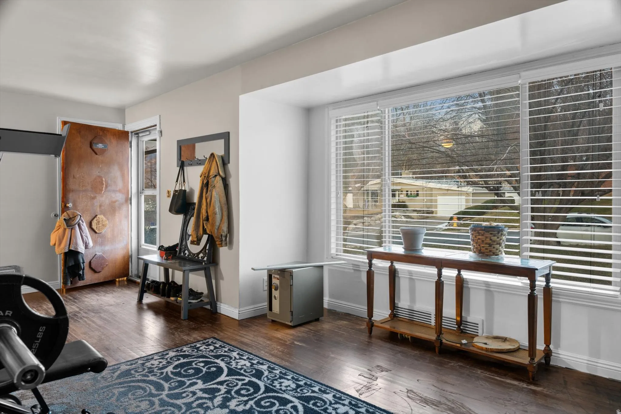 Spacious room featuring wood-finish flooring, a large bay window with horizontal blinds, and a solid wood entry door