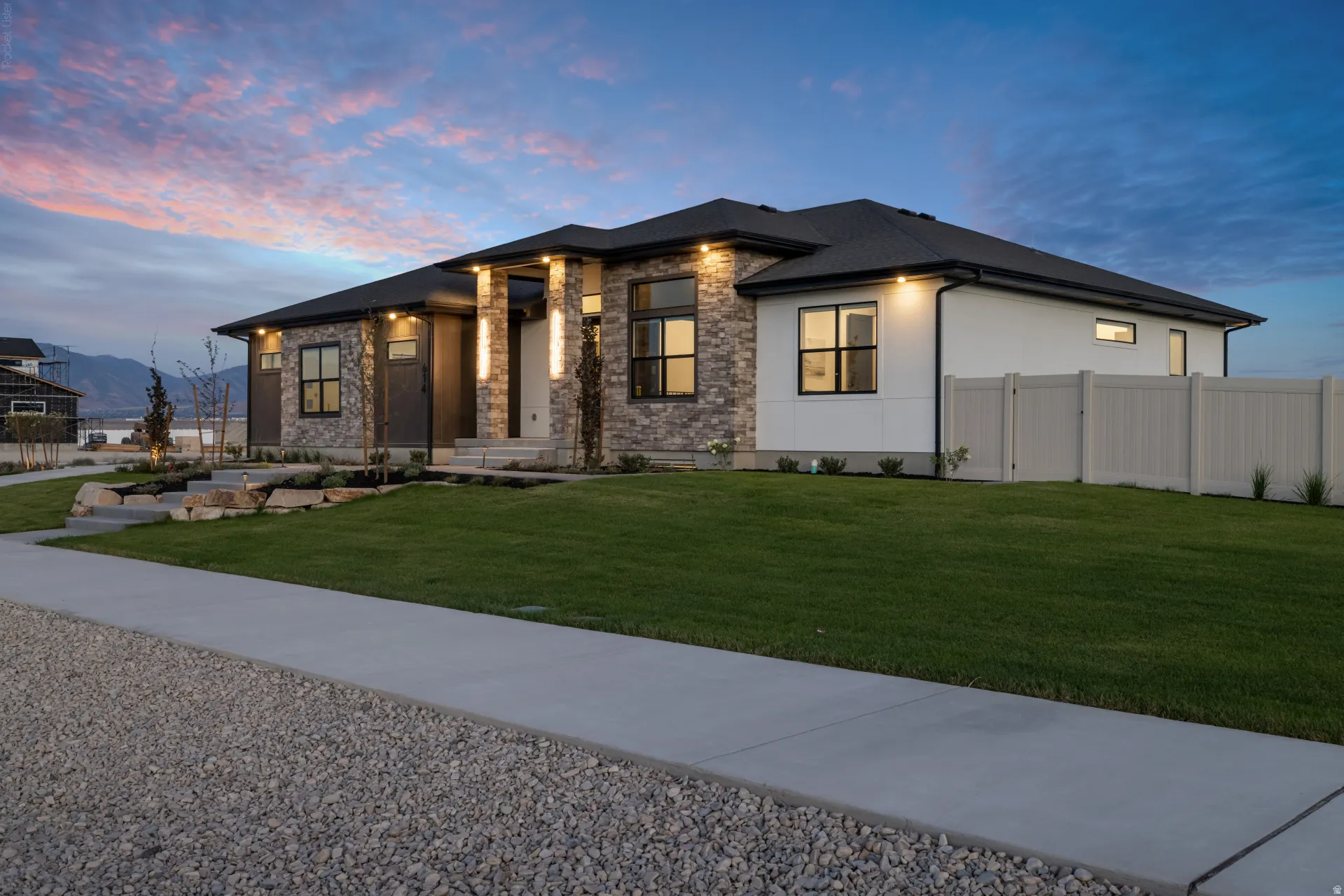 Modern ranch-style residence featuring a stone and stucco facade, black-framed windows, a dark shingled roof, a manicured lawn, and a light-colored privacy fence
