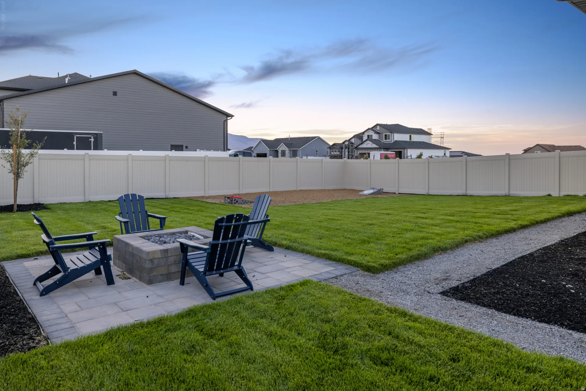 Expansive backyard featuring a paver patio with a built-in stone fire pit, a manicured lawn, gravel pathways, and a privacy fence