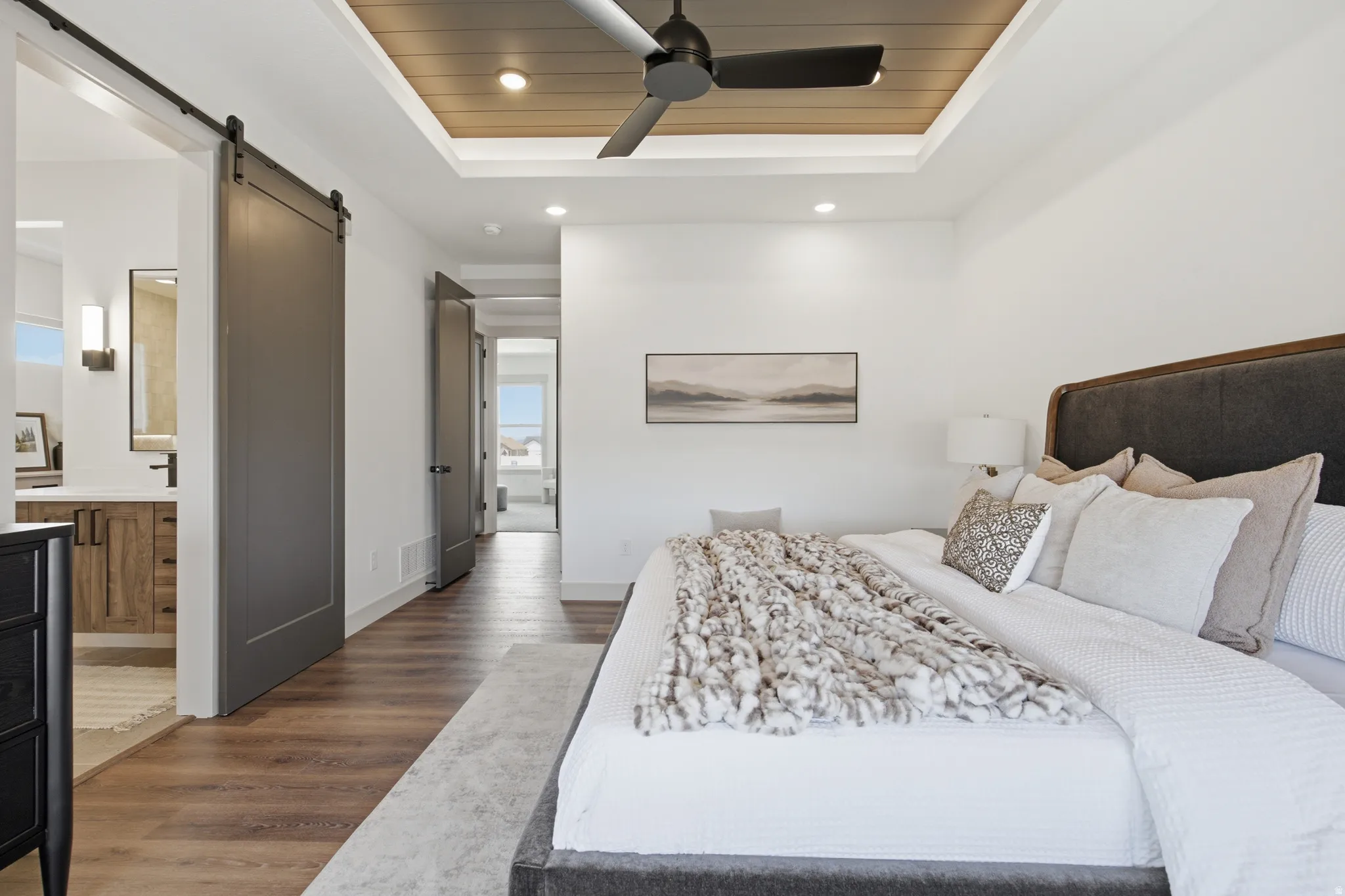 Spacious bedroom featuring wood-finish flooring, a recessed tray ceiling with wood paneling and ceiling fan, and a wide entryway with a modern barn door