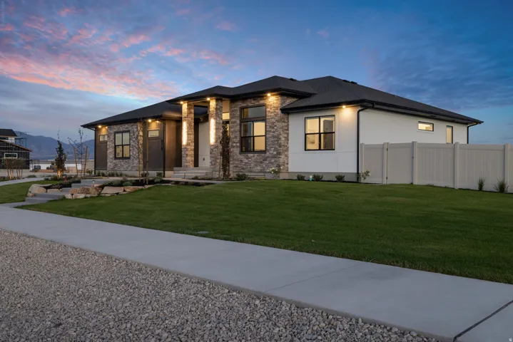 Modern ranch-style residence featuring a stone and stucco facade, black-framed windows, a dark shingled roof, a manicured lawn, and a light-colored privacy fence