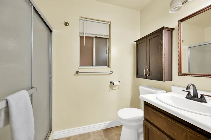 Bright and updated bathroom featuring a dual vanity with white countertop and wood-finish cabinetry, framed mirror with decorative surround, single-handle faucet, ceramic tile flooring, toilet, and glass-enclosed shower stall with towel bar
