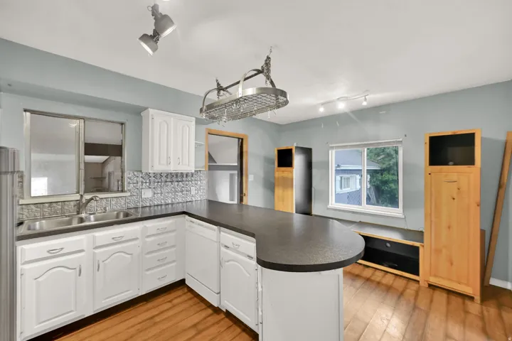 White cabinetry with brushed nickel hardware, stainless steel double basin sink, gray textured backsplash, and dark countertop with an integrated breakfast bar