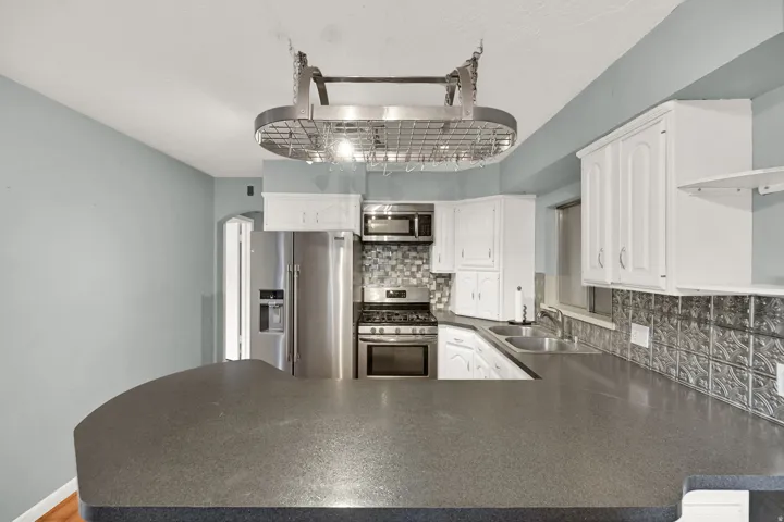 Curved countertop with integrated dual sinks, stainless steel appliances, white cabinetry, decorative tile backsplash, and overhead pot rack