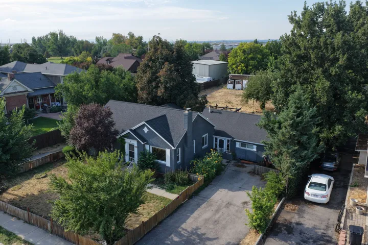Charming single-story residence featuring a dark-finished exterior, established landscaping with mature trees throughout the grounds, a paved driveway with attached carport, and a fenced perimeter