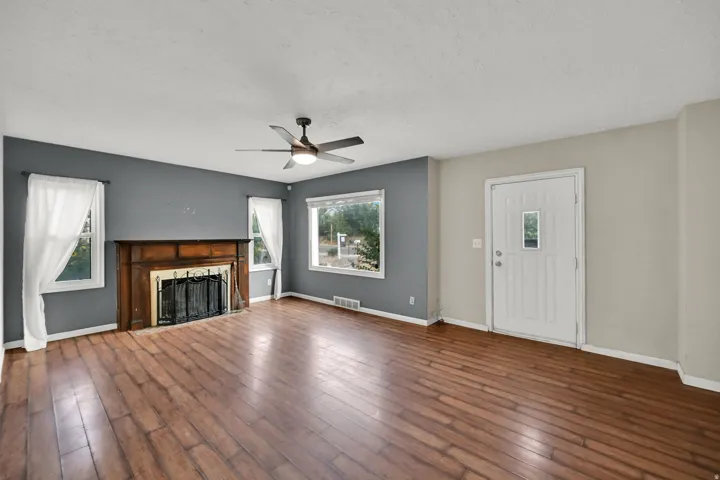 Wood-finish flooring throughout, a decorative fireplace with an ornate wood mantel, multiple windows, and a ceiling fan with integrated lighting