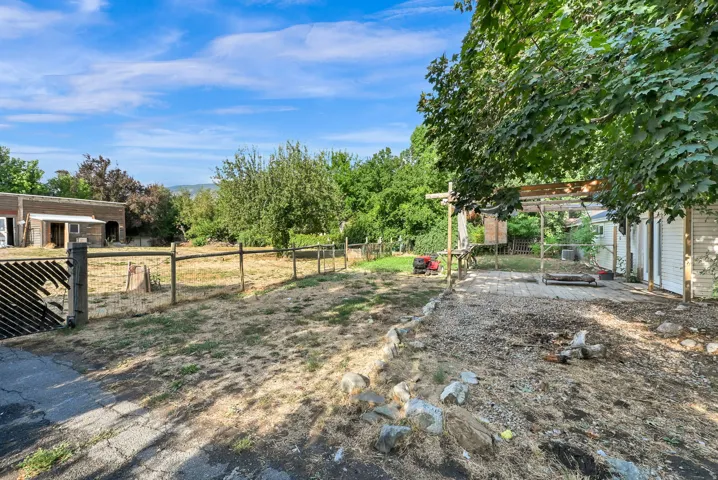 Expansive outdoor space featuring a paved patio area, wood fencing, mature trees, and a distant building structure